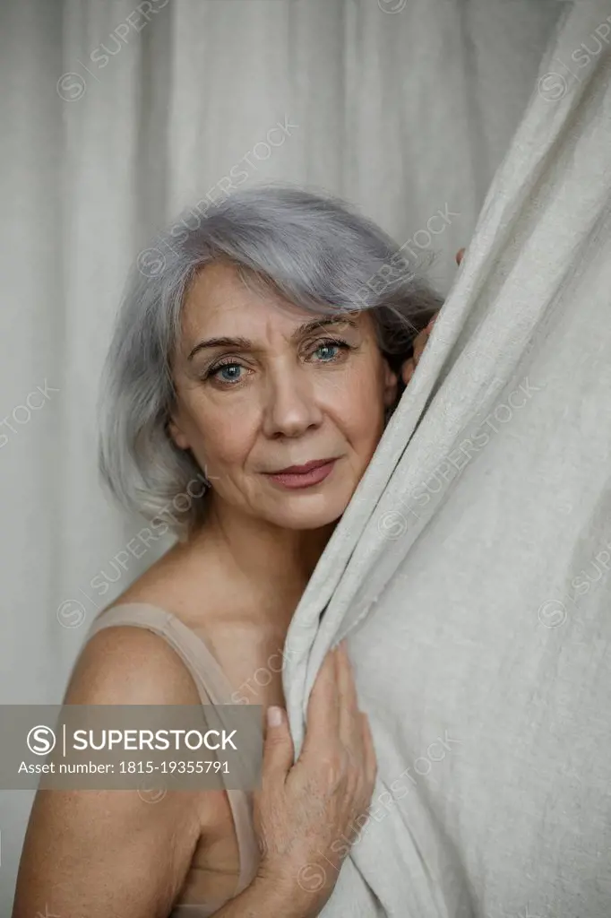 Senior gray haired woman by curtain at home