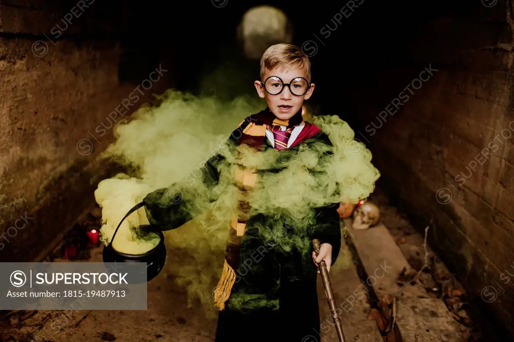 Cute boy with cauldron and magic wand standing amidst yellow smoke in tunnel
