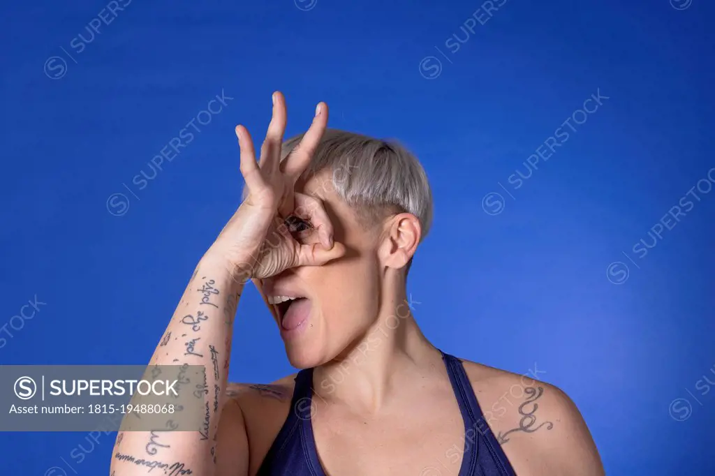 Woman with short hair looking through fingers against blue background