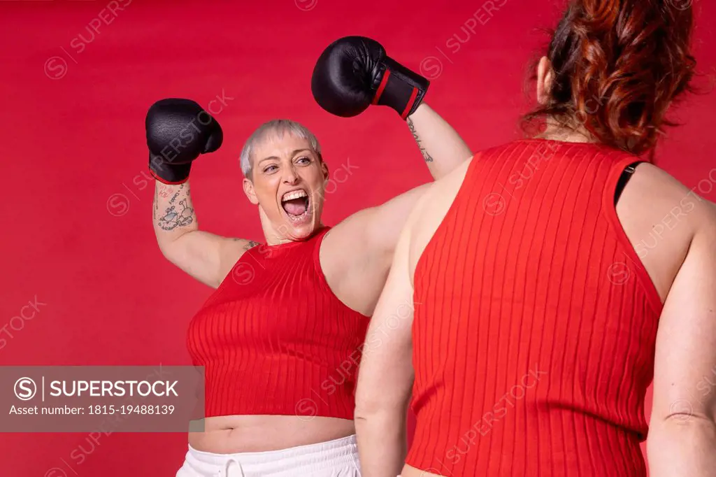 Woman wearing boxing gloves flexing muscles looking at friend against red background