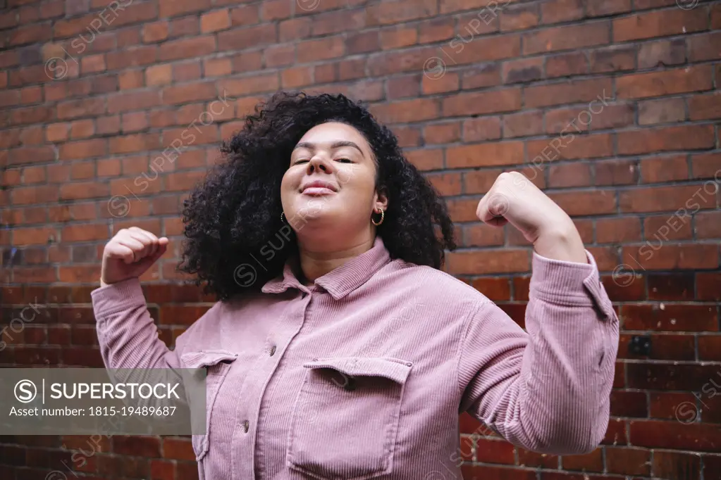 Confident young woman with curly hair flexing muscles in front of brick wall