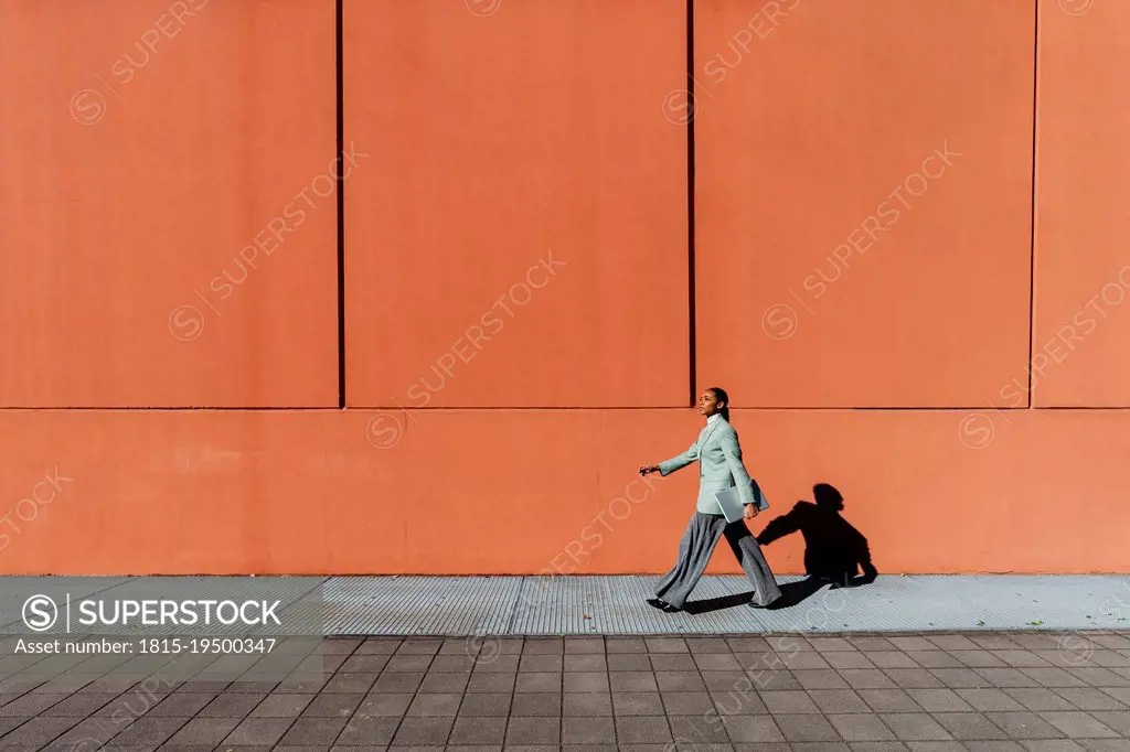 Young businesswoman with laptop walking by orange wall