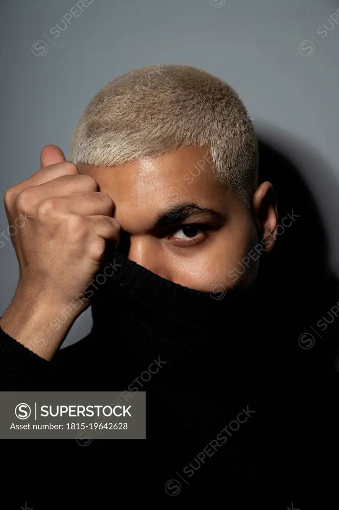 Young man covering face with turtleneck top against gray background