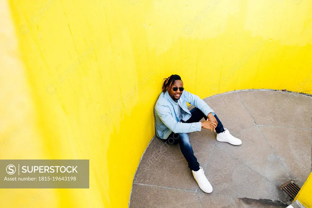 Young man with sunglasses sitting in front of yellow wall