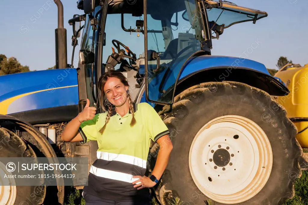 Happy female farmer gesturing thumbs up in front of tractor on sunny day