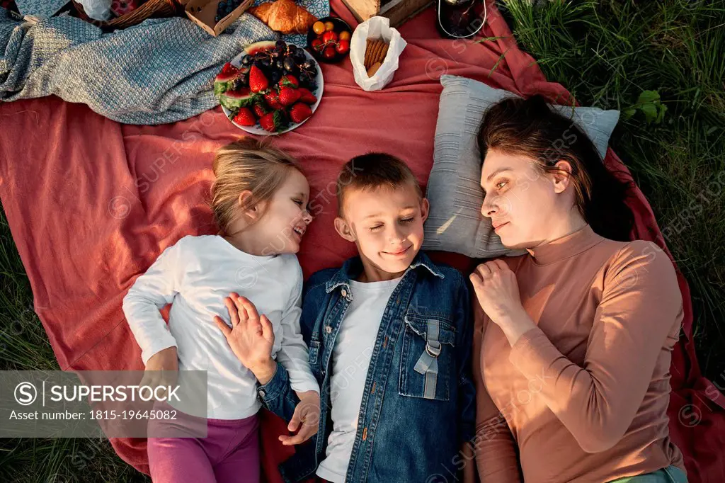 Mother looking at children lying on picnic blanket in field