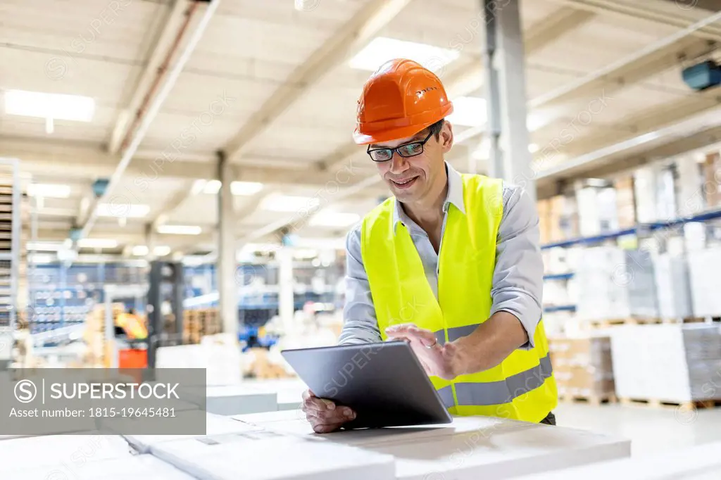 Smiling worker using tablet PC leaning on box in warehouse