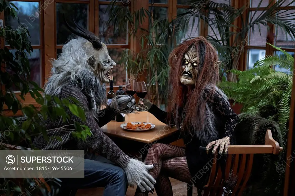 Man and woman in spooky costume toasting wine glasses sitting at table