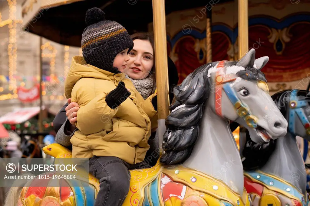 Woman supporting boy sitting on carousel horse in Christmas market