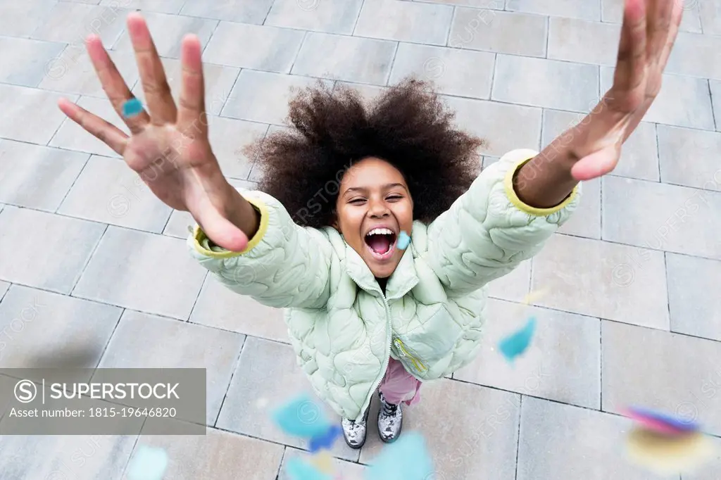Cheerful girl with arms raised standing amidst confetti falling on footpath
