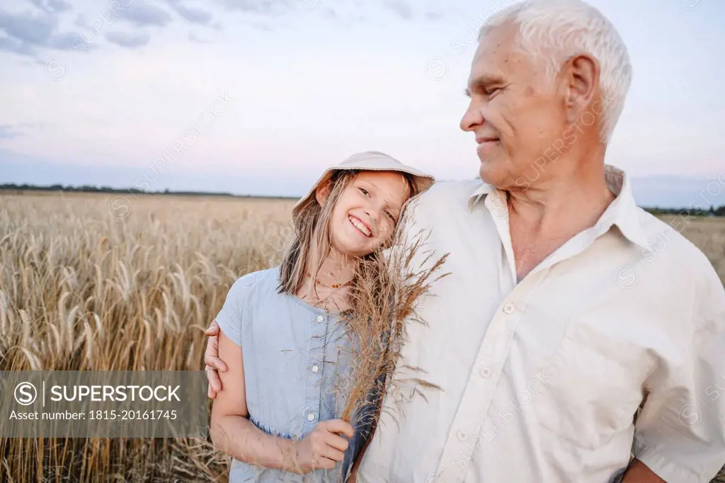 Smiling girl holding crops with grandfather standing at rye field