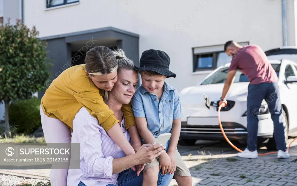 Mother and children checking smartphone while father is charging electric car