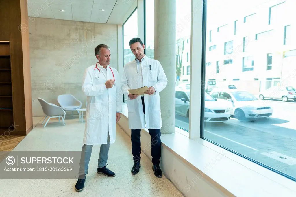 Doctors in lab coats discussing over medical record standing by window at hospital