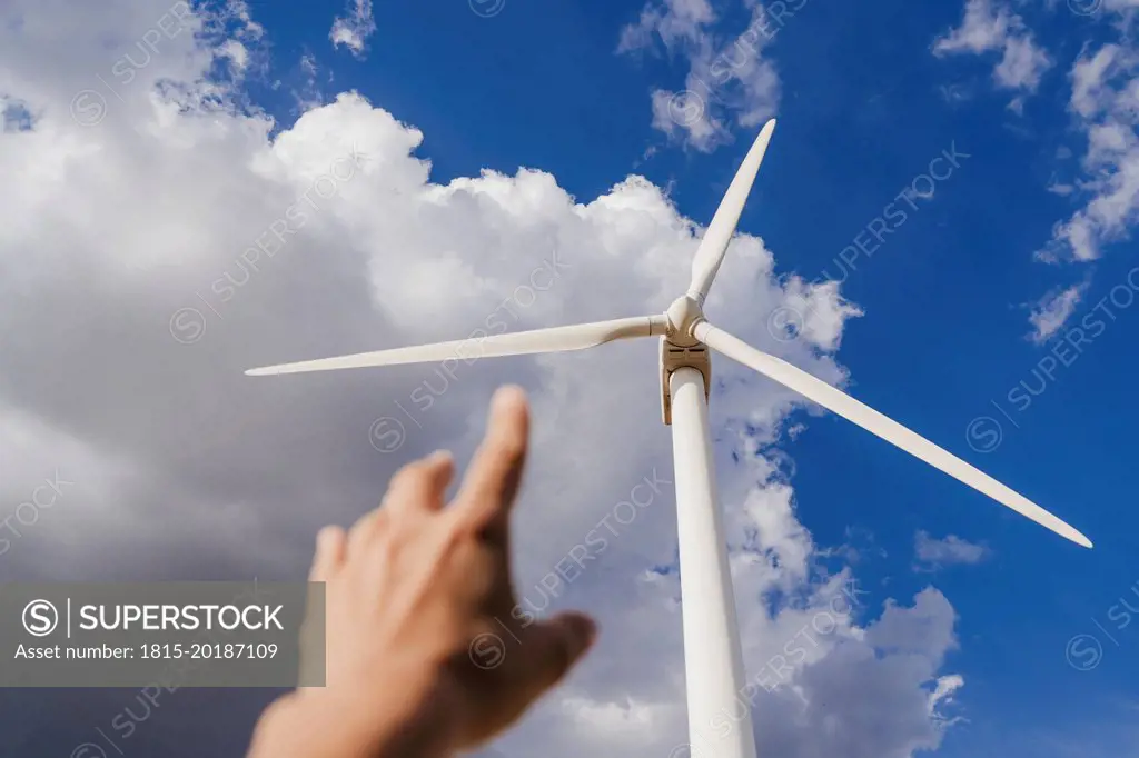 Hand of woman gesturing at rotor of wind turbine below clouds in sky