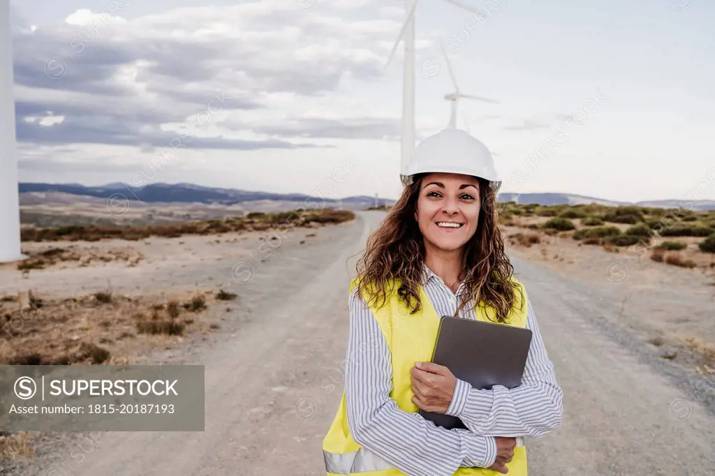 Smiling engineer wearing hardhat holding laptop at wind farm