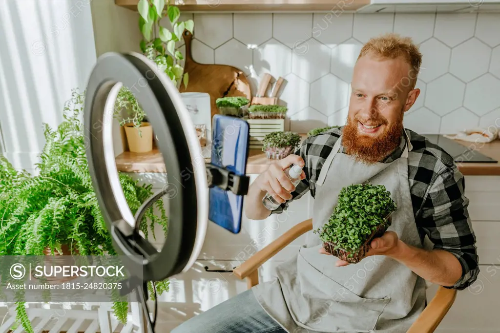 Smiling man spraying water on microgreens sitting in front of smart phone