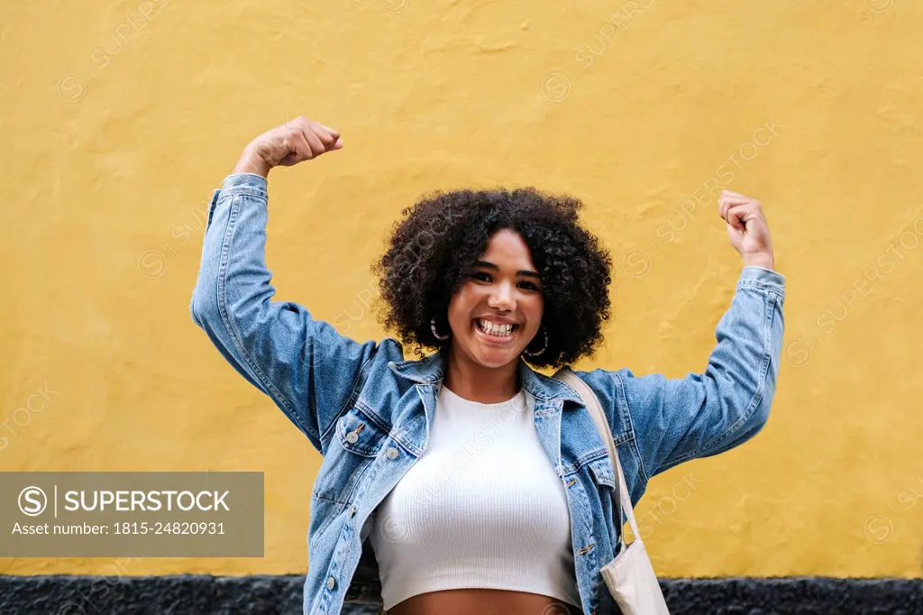Cheerful young woman flexing muscles in front of wall