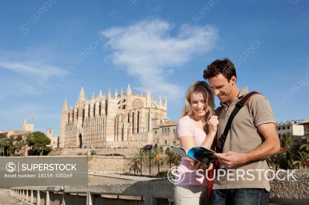 Spain, Mallorca, Palma, Couple looking in guide book with St Maria Cathedral in background, smiling
