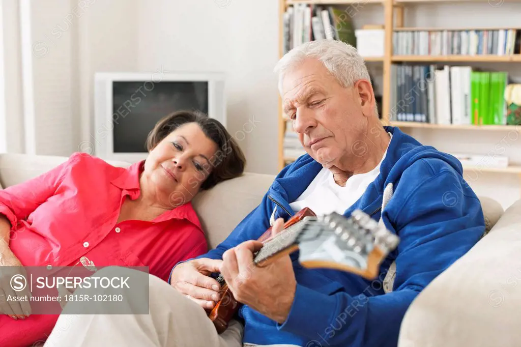 Germany, Leipzig, Senior man playing electric guitar on sofa, woman sitting beside