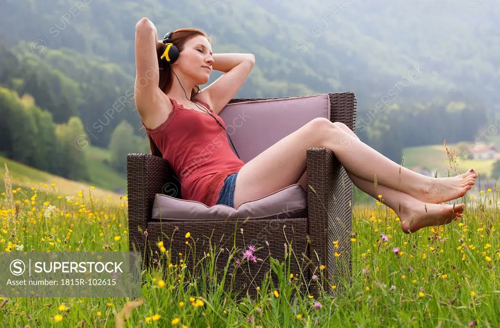 Austria, Young woman sitting on sofa and listening music in field of flowers