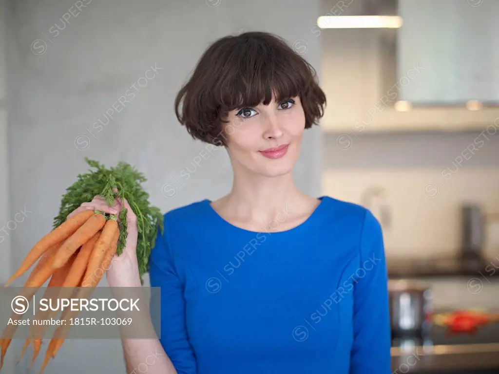 Germany, Cologne, Young woman with carrots in kitchen, smiling, portrait
