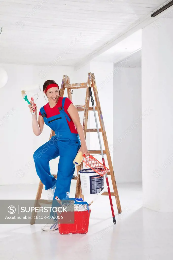 Germany, Bavaria, Young woman leaning on step ladder with paint equipments, smiling, portrait