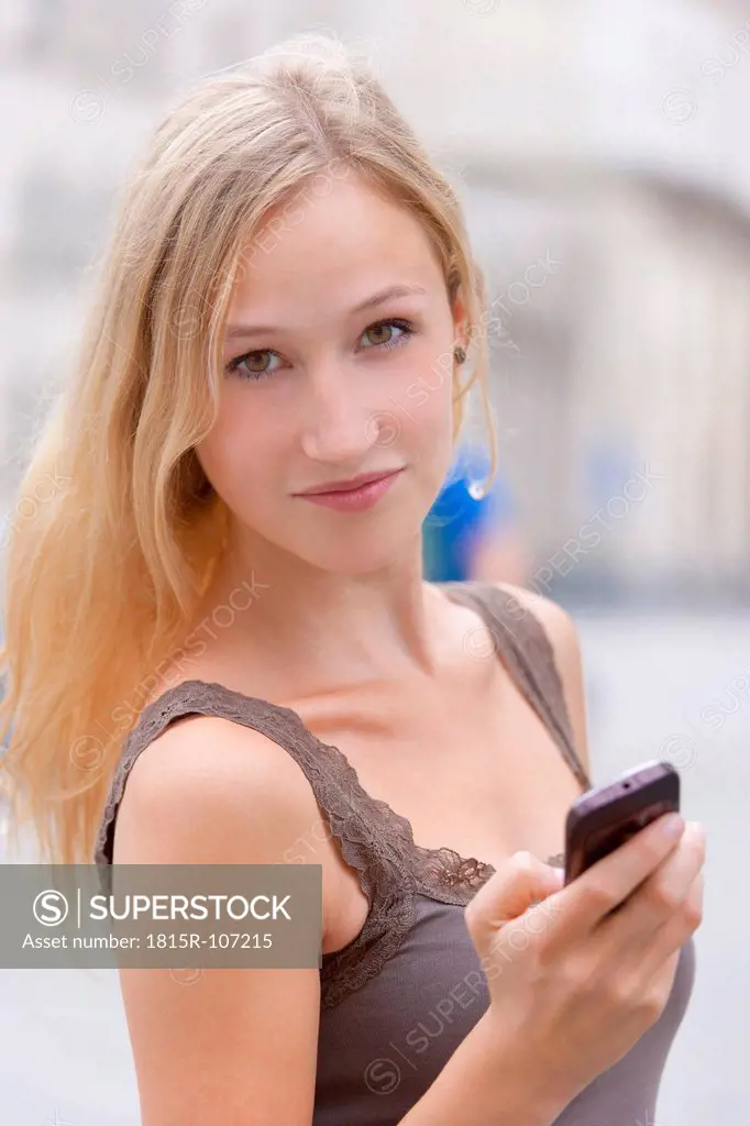 Germany, Bavaria, Munich, Young woman standing with smart phone in front of Ludwig Maximilian University