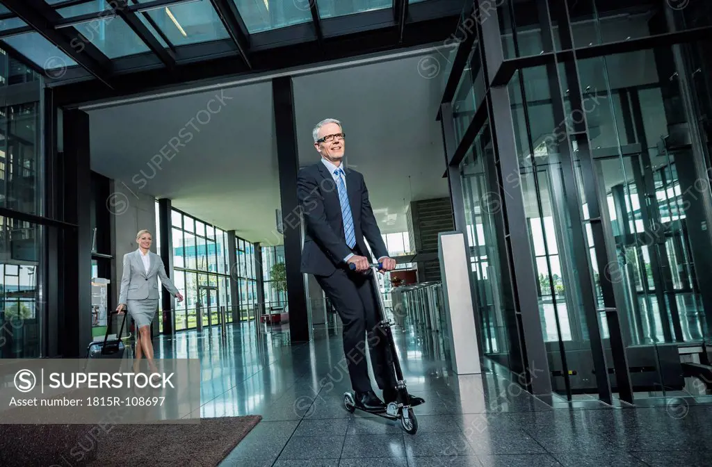 Germany, Stuttgart, Businesswoman with wheeled luggage, man riding scooter at office building
