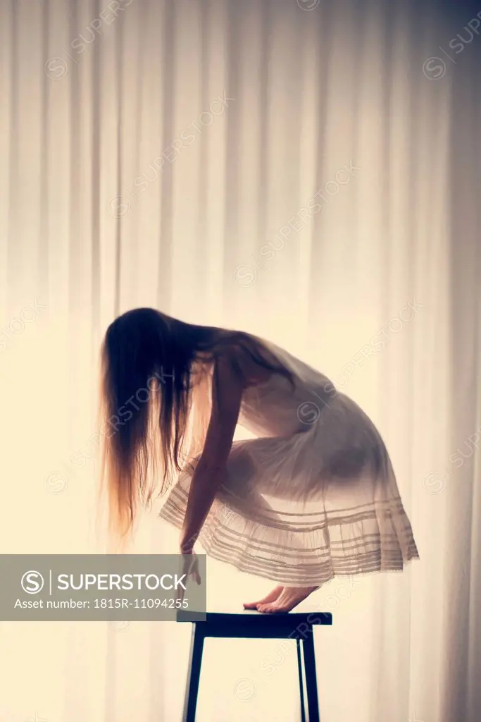 Young woman crouching on a stool in front of a white curtain, back view