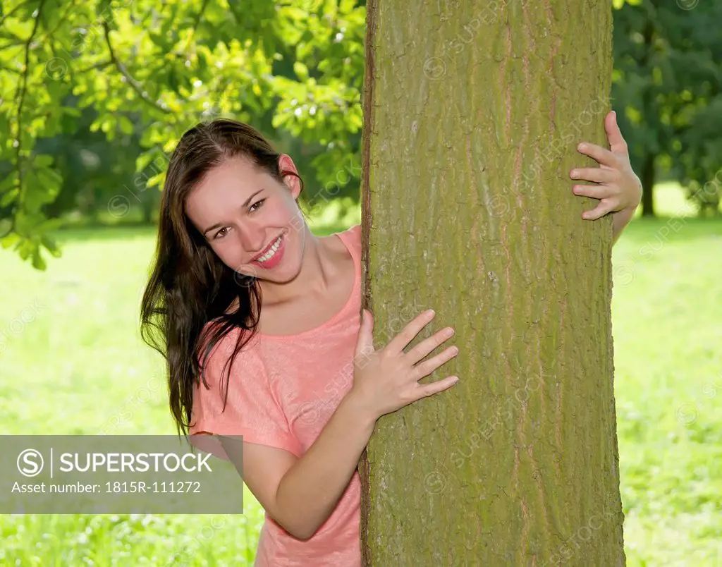 Germany, Berlin, Young woman embracing tree trunk, smiling, portrait