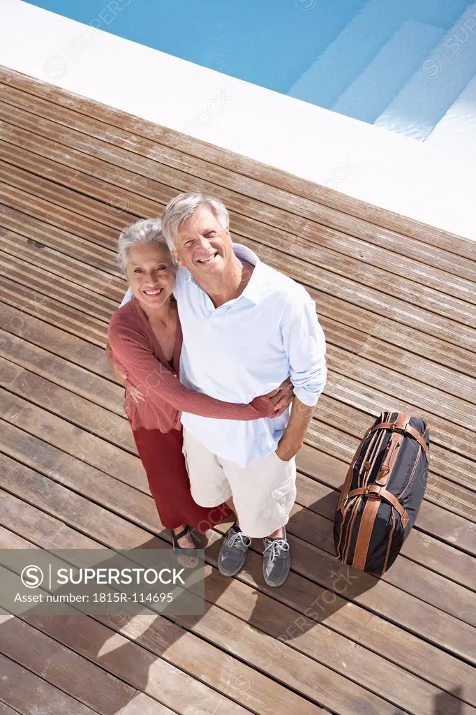 Spain, Senior couple standing with suitcase at swimming pool, smiling, portrait