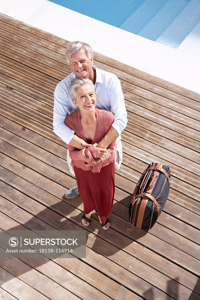 Spain, Senior couple standing with suitcase at swimming pool, smiling, portrait