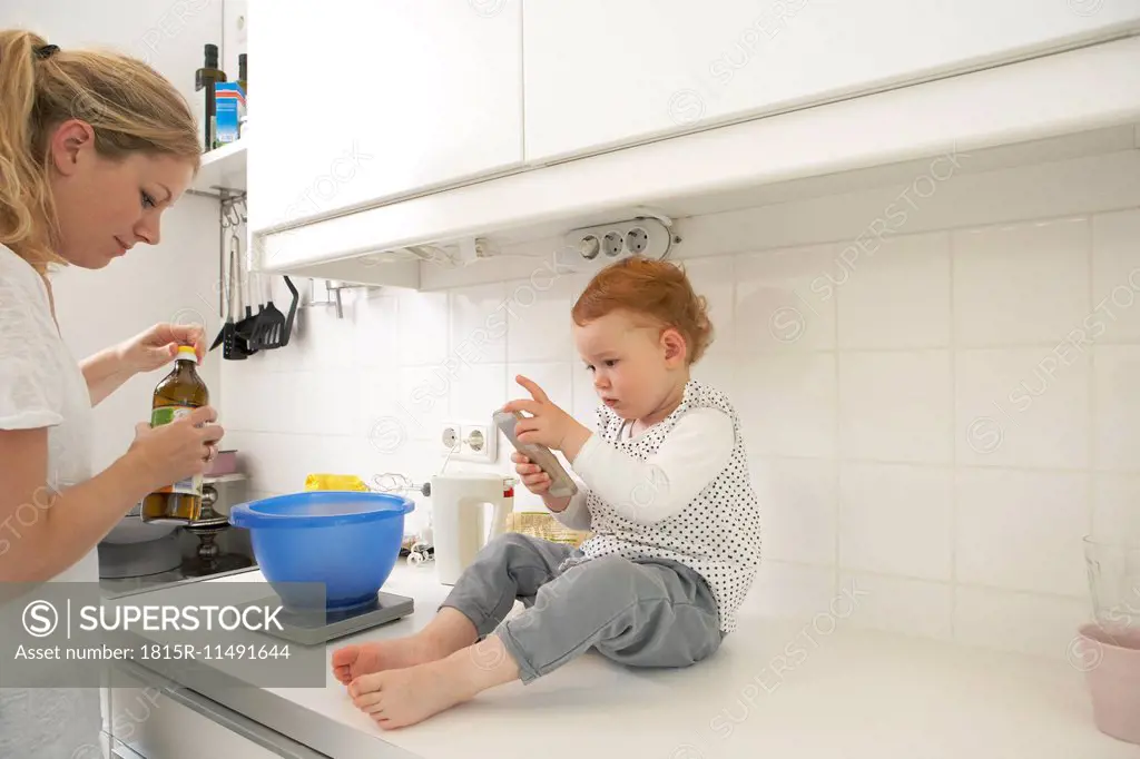 Little girl sitting on kitchen counter with smartphone while her mother baking