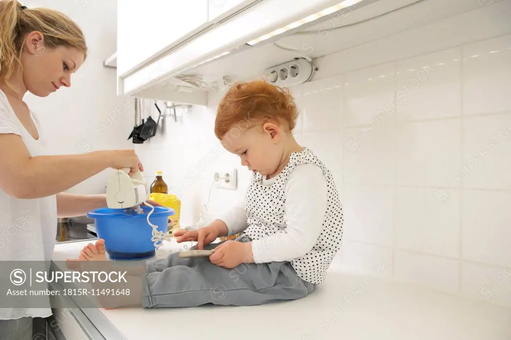 Little girl sitting on kitchen counter with smartphone while her mother baking