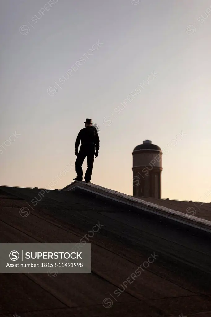 Germany, silhouette of chimney sweep standing on rooftop at twilight
