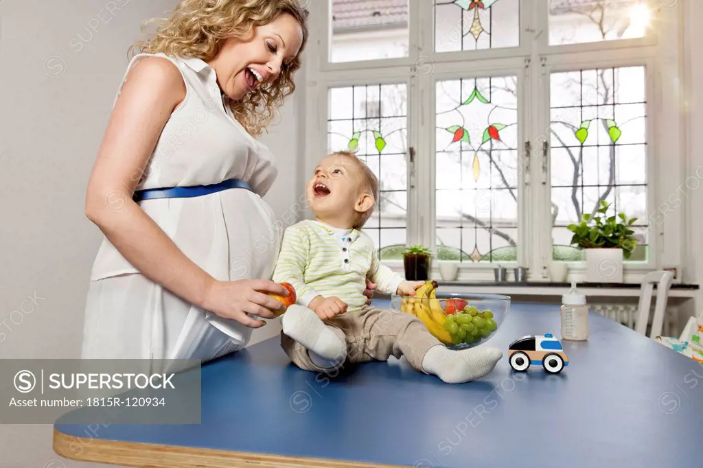 Germany, Bonn, Pregnant mother showing fruits to son in living room, smiling