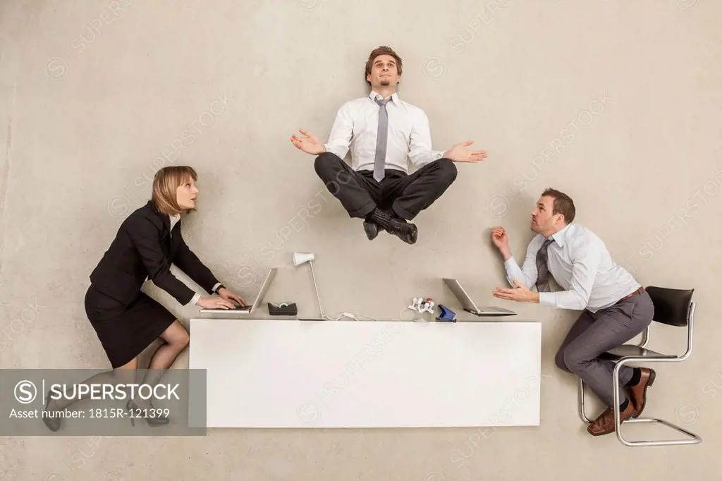 Businessman meditating above office desk while others working