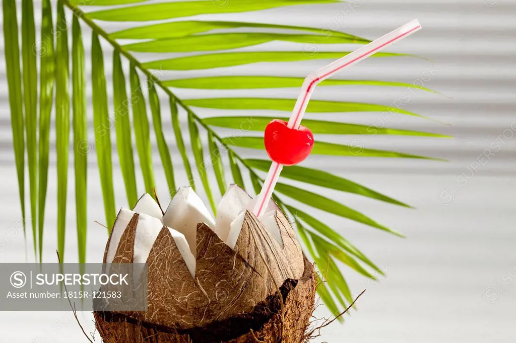 Coconut with drinking straw, cherry and palm leaf in background, close up