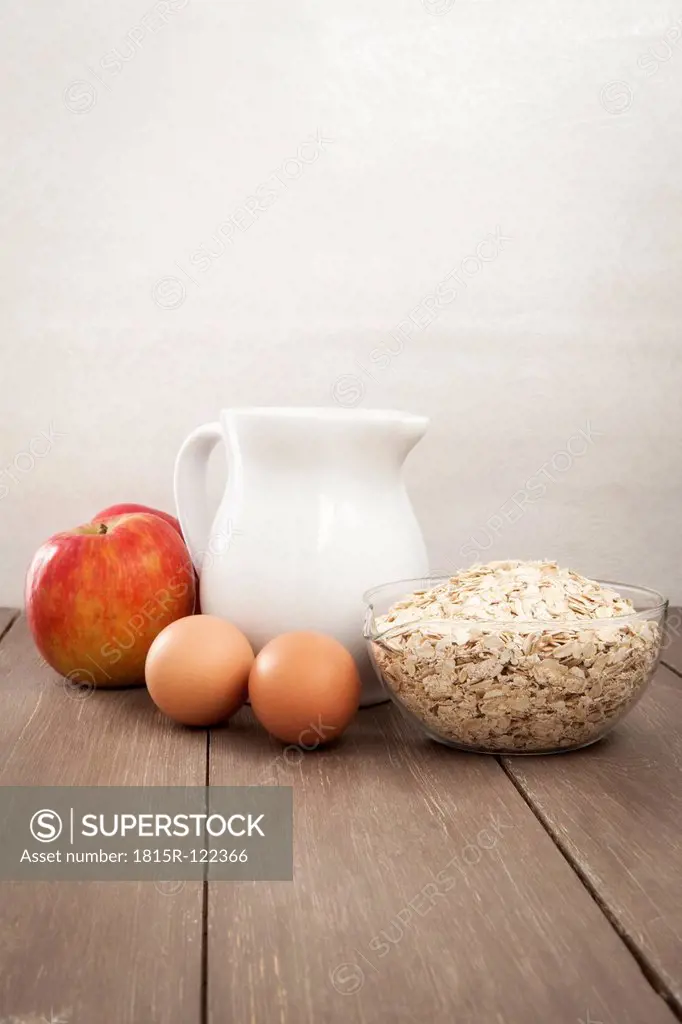 Milk jar with apples, eggs and bowl of oat flakes on wooden table, close up