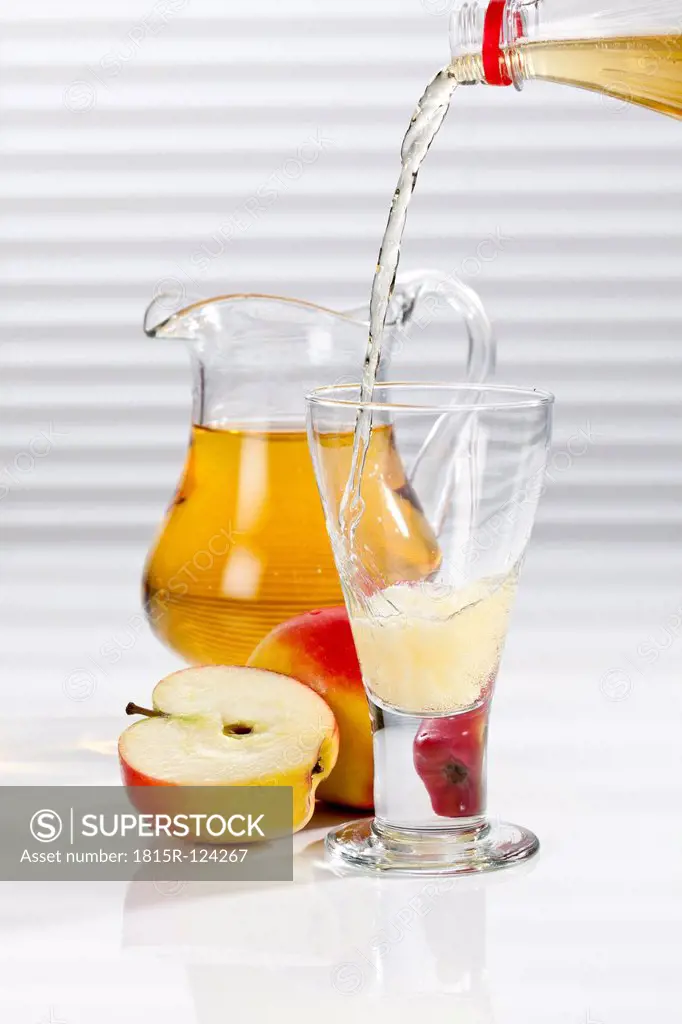 Apple juice being poured into glass besides apples and pitcher