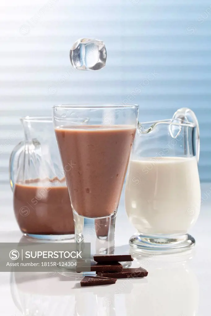 Ice cube falling into Glass of chocolate milk with carafes and pieces of chocolate besides
