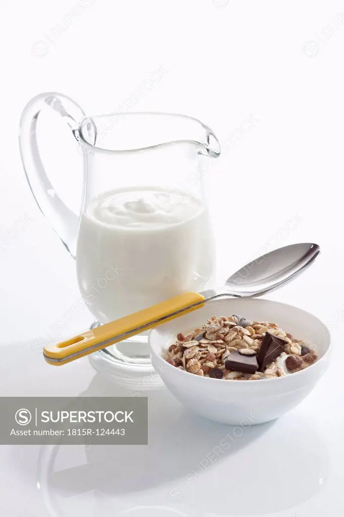 Bowl of muesli yogurt with chocolate beside yogurt carafe on white background, close up