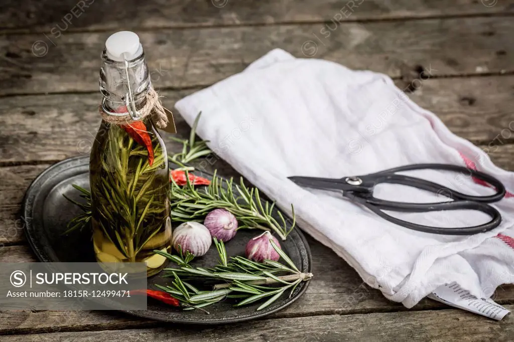 Rosemary oil in bottle, garlic, rosmary and chilli peppers on plate, scissors and kitchen towel on wood