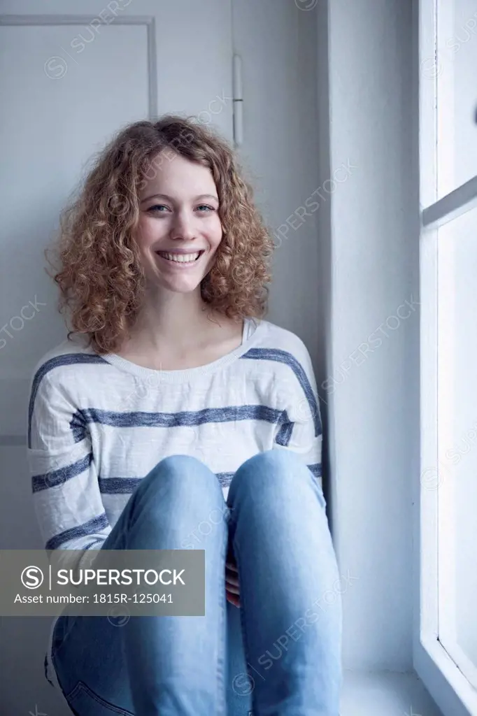 Germany, Bavaria, Munich, Young woman sitting at window, smiling, portrait
