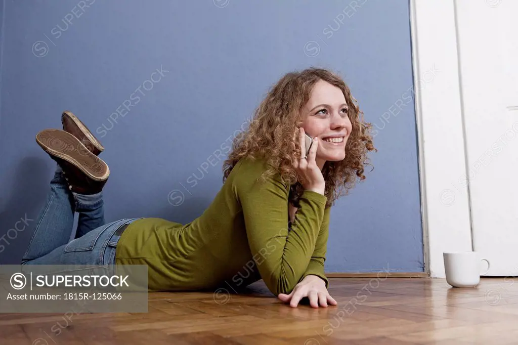 Germany, Bavaria, Munich, Young woman lying on floor and talking on cell phone, smiling