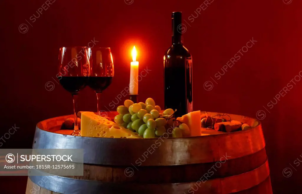 Two glasses of red wine, bottle, grapes, cheese and lighted candle on a wine cask in a wine cellar