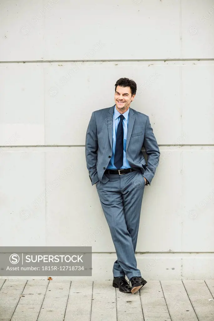 Portrait of smiling businessman wearing grey suit leaning against concrete wall