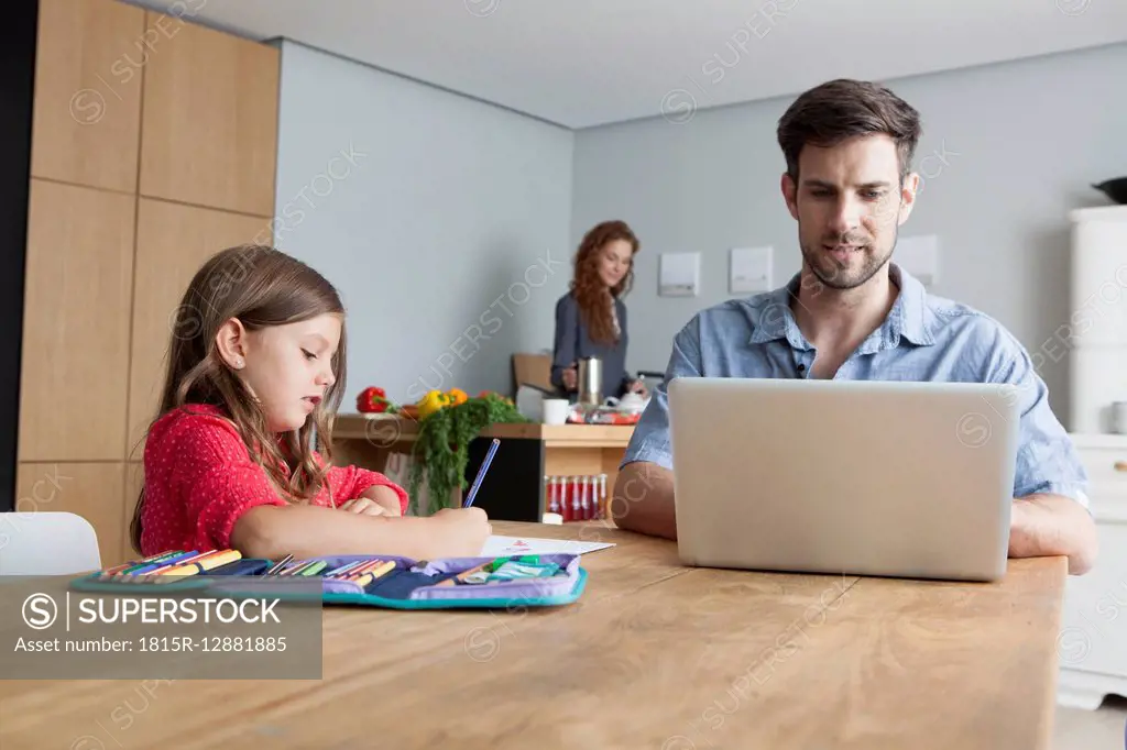 Man sitting at kitchen table using laptop while his little daughter painting