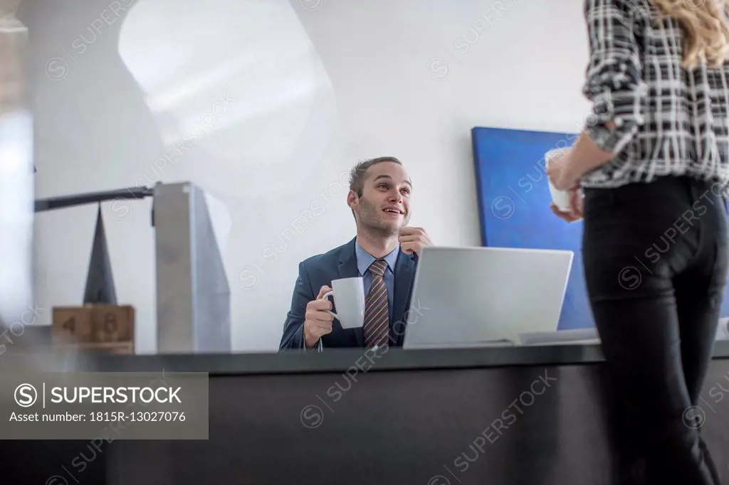 Businessman at desk with cup of coffee looking at colleague