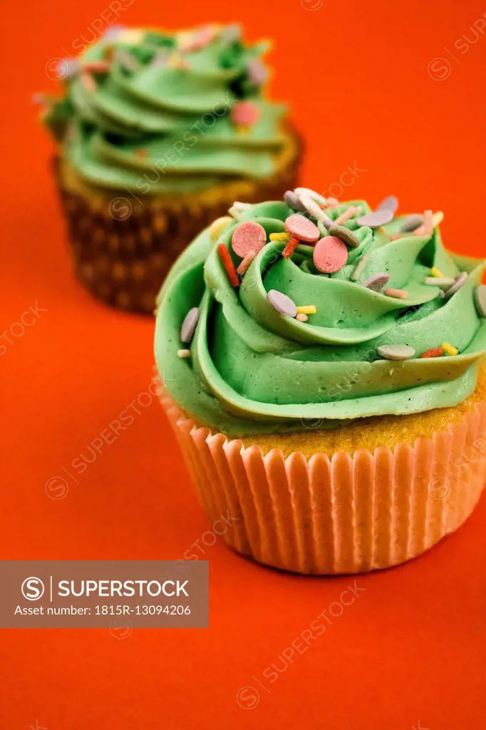 Two cupcakes with green cream and baking dekor in front of red background